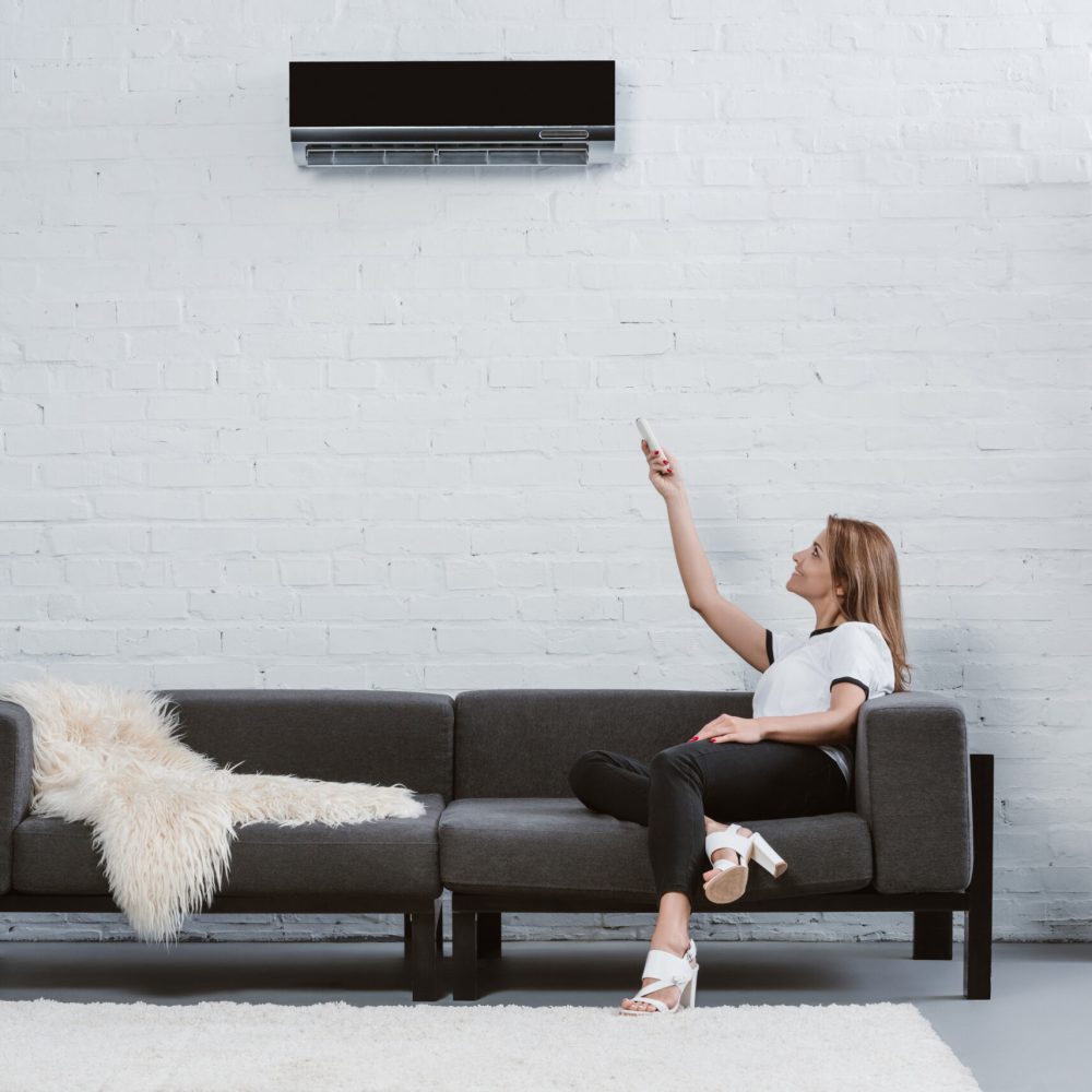 beautiful young woman sitting on sofa and pointing at air conditioner hanging on wall with remote